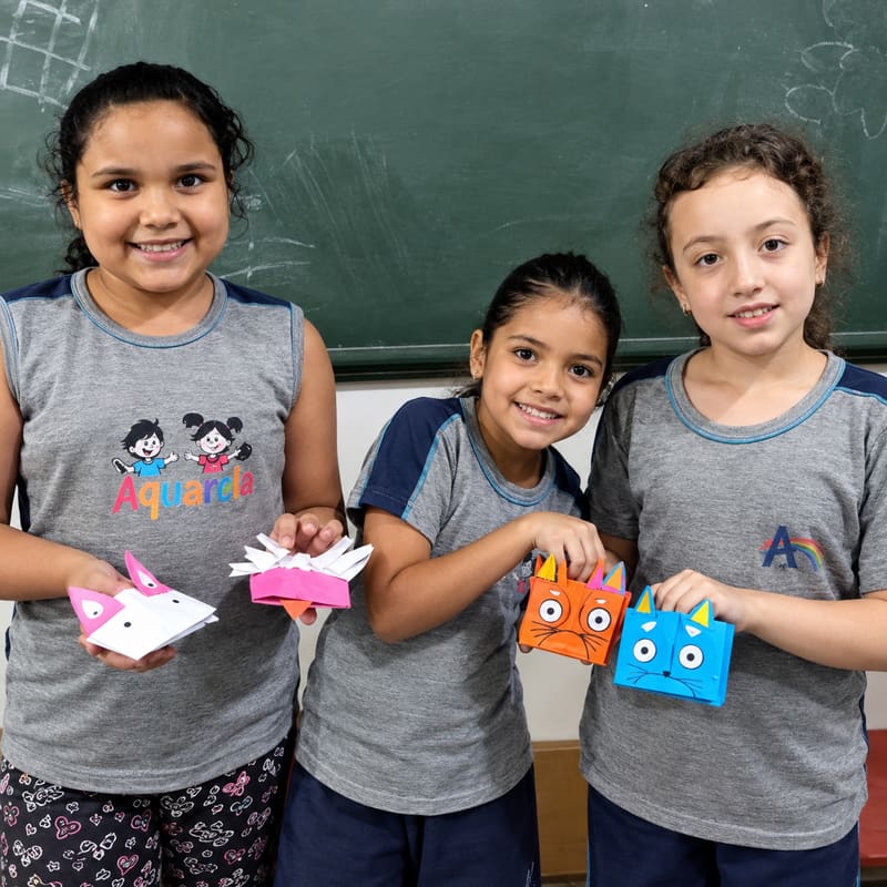 Três alunas de uniforme, em frente ao quadro-negro, mostram as dobraduras de papel colorido que fizeram em sala