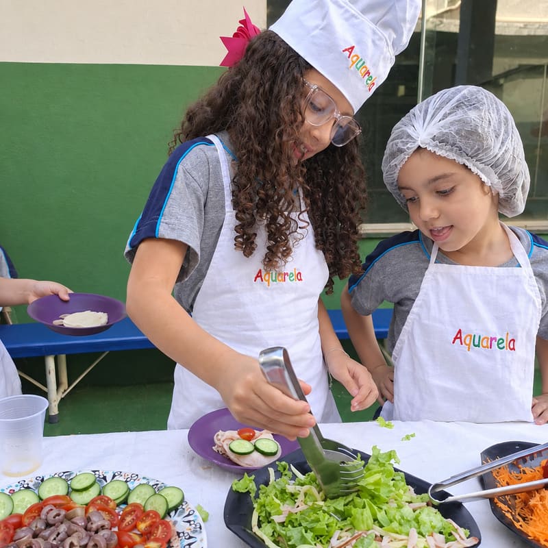 Duas alunas de avental Aquarela, uma de touca de chef e outra de touca descartável, servem salada com pegador; na mesa, pratos com tomate, pepino e cenoura ralada