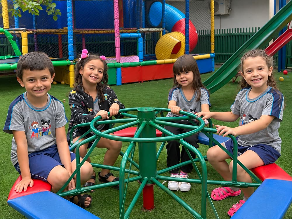 Quatro crianças de uniforme sentadas no gira-gira do playground, com o brinquedão colorido e o escorregador ao fundo