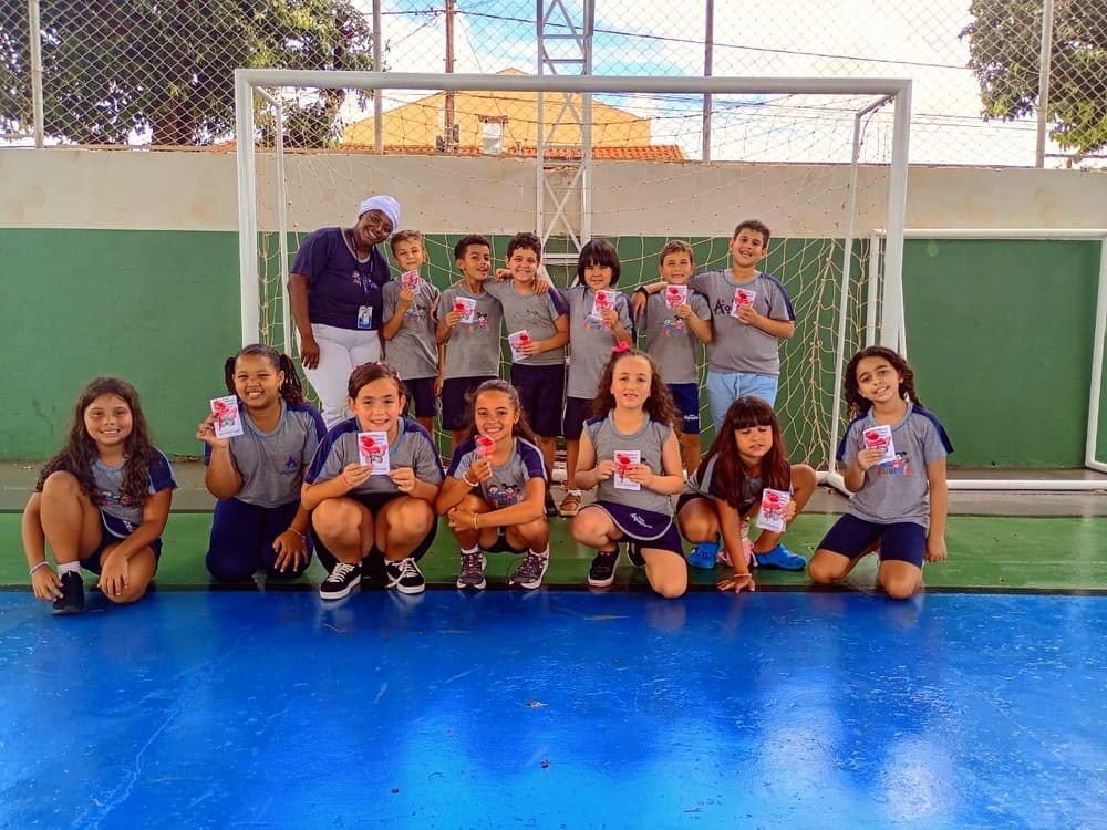 Turma de alunos de uniforme posa com a professora em frente ao gol da quadra da escola