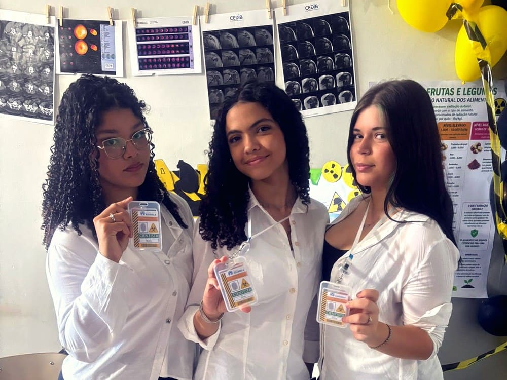Três alunas de jaleco branco mostram seus crachás em uma feira de ciências, com radiografias e cartazes na parede
