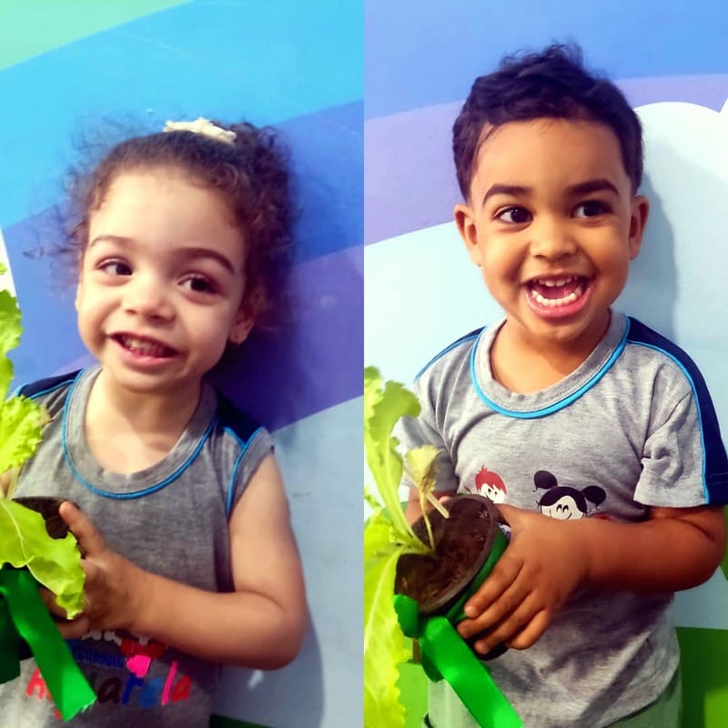Duas crianças de uniforme sorriem segurando mudas de alface em vasinhos