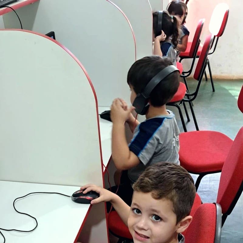 Dois meninos em cabines de computador: um usa fones de ouvido e o mouse, o outro sorri para a câmera