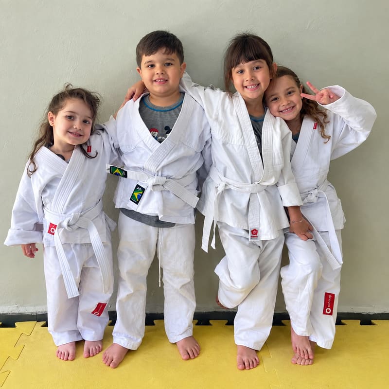 Quatro crianças de quimono e faixa branca, abraçadas e sorrindo, descalças sobre o tatame amarelo